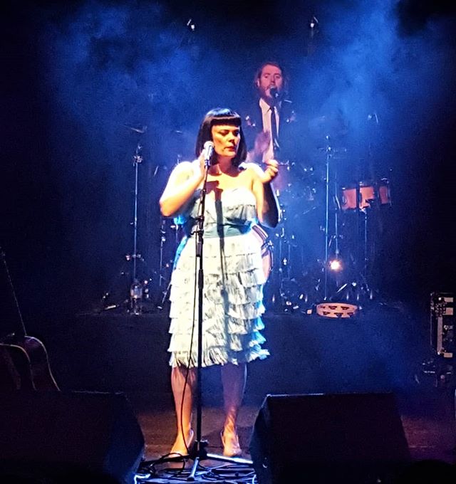 a female singer wearing a very pale blue dress at the microphone singing. The background is an inky blue and you can just see one of the band members in the haze of the lights.