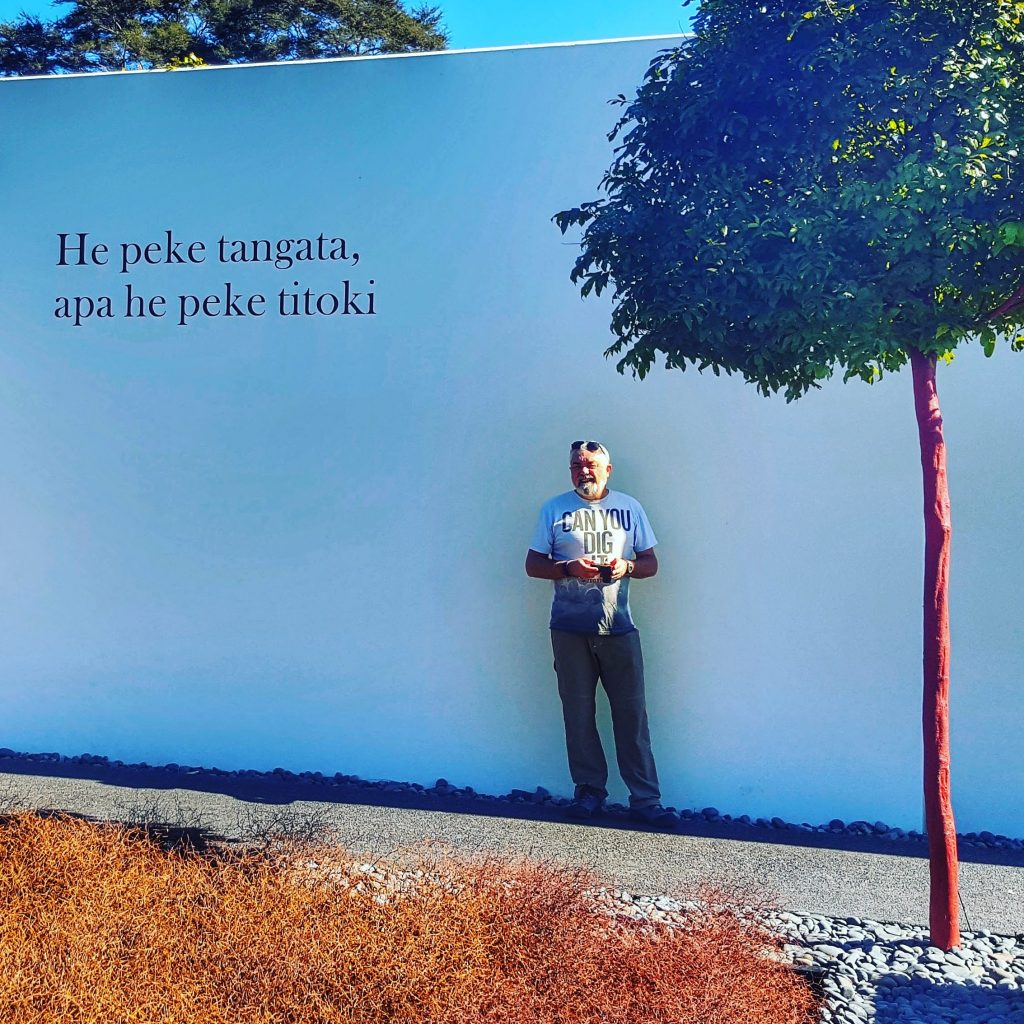 man standing against a white wall with the words He peke tangata, apa he peke titoki’ written on it. That means "The human family lives on while the branch of the titoki falls and decays.